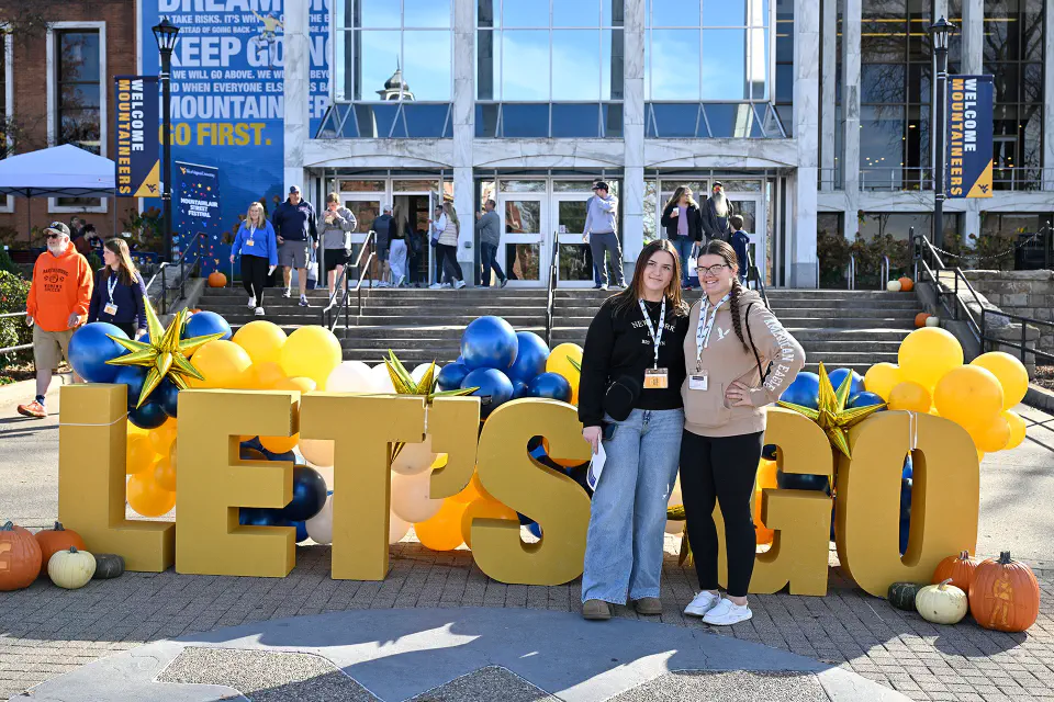 Two prospective students pose for a photo in front of the big Let's Go letters decorated with gold and blue balloons. The Mountainlair Student Union is in the background.